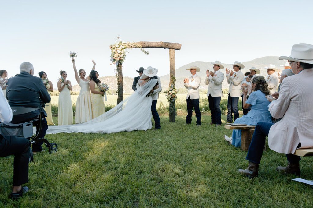First kiss at a wedding ceremony with the bridal party cheering on the couple after saying I do. 